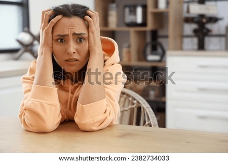 Beautiful young afraid woman sitting at table in kitchen Royalty-Free Stock Photo #2382734033