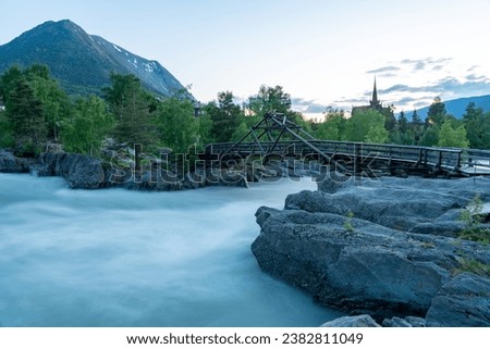 Bridge crossing a  big river flowing through the Lom village. Image taken during summer night. Slow shutter speed long exposure image. Old wooden church on the background. River through the cliffs