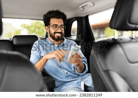 Cheerful handsome millennial indian guy in casual wearing eyeglasses sitting on auto back seat, using smartphone and smiling, hindu man chatting with friends while sitting in taxi Royalty-Free Stock Photo #2382811249