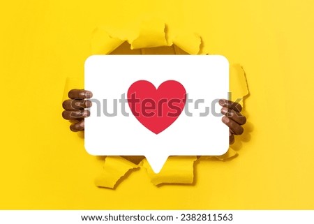 Closeup of african american guy holding and showing social media message icon with heart symbol, liking photo through hole in yellow paper background. Online communication and SMM. Collage