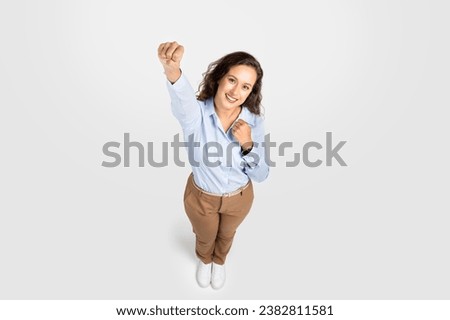 Smiling confident young woman playfully, embodying iconic super hero flight pose. Her arm extends powerfully forward, fist clenched, as she emanates lively, imaginative spirit on gray background Royalty-Free Stock Photo #2382811581