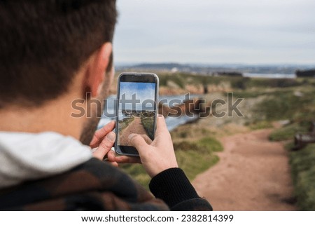 Rear view of a young man photographing the landscape through his mobile phone while standing on a cliff against a cloudy sky.