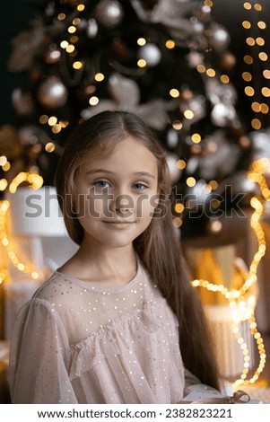 Cute little girl with long hair in dress sitting on the floor and opens a box with a present for background Christmas tree. Xmas holiday concept