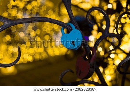 Blue metal love lock in shape of heart, as a symbol of an eternal relationship, hangs on a metal tree against a background of bright lights in the park in the evening.