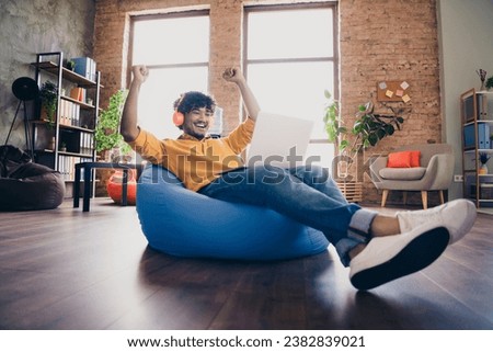 Photo portrait of nice young male wear yellow shirt sit blue bean bag raise fists celebrate done work interior loft industrial style