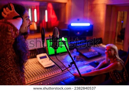 Profile of a male singer singing at the microphone and recording a songs in a recording studio. An artist is making audio material for a single. In a blurry background is a technician recording.