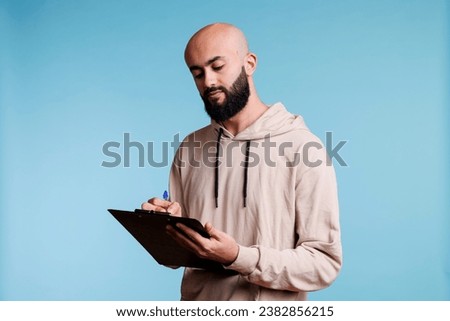 Young arab man writing in clipboard with concentrated facial expression. Focused arabian bald bearded person making list and taking notes while standing on blue background