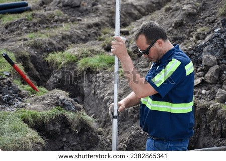 Male construction worker wearing navy blue with high visibility lines, working in dirt hole, holding a measuring stick, in the summer. Royalty-Free Stock Photo #2382865341