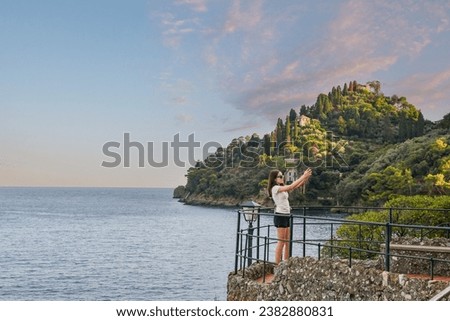Teenage girl (13 years old) taking a selfie with her smarphone on the Terrace of Punta Caieca viewpoint, with the Promontory of Portofino in the background, at sunset in summer, Genoa, Liguria, Italy Royalty-Free Stock Photo #2382880831