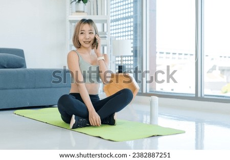 Asian young female athlete teenager in sportswear sport bra sitting smiling on yoga pilates mat holding using smartphone in hand in gym after training exercising working out.