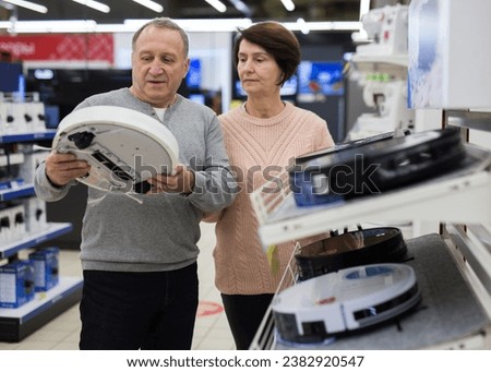 Middle aged wife and husband picking robotic vacuum cleaner in shop of household appliances.