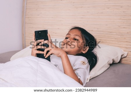 Cute little kid girl watching video on smartphone with smiley face alone on the bed.