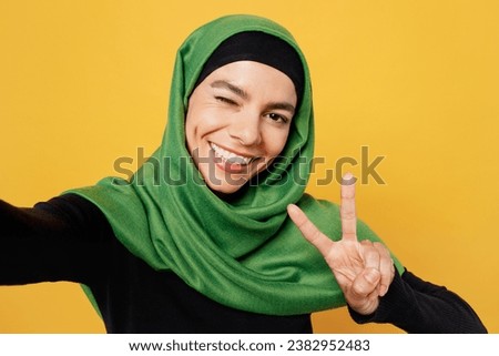 Close up young asian muslim woman wears green hijab abaya black clothes do selfie shot pov mobile cell phone wink isolated on plain yellow background People uae middle eastern islam religious concept