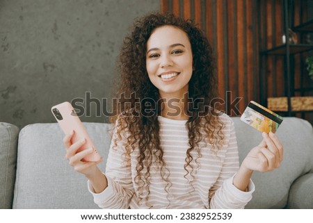 Cheerful young happy woman wear casual clothes sits on grey sofa couch using mobile cell phone credit bank card shopping online stay at home hotel flat rest relax spend free time in living room indoor