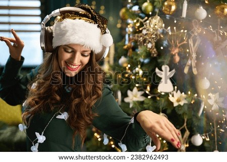 Christmas time. smiling modern female with Santa hat in green dress listening to the music with headphones and dancing near Christmas tree in the modern house. Royalty-Free Stock Photo #2382962493