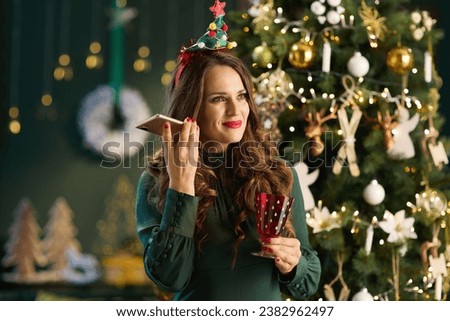 Christmas time. smiling modern woman with glass in green dress talking on a smartphone near Christmas tree in the modern house. Royalty-Free Stock Photo #2382962497
