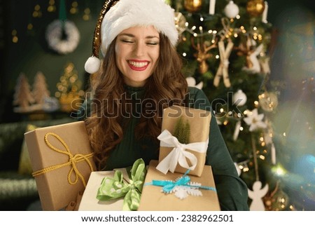 Christmas time. happy stylish woman with Santa hat and eco packaged present boxes in green dress near Christmas tree at modern home. Royalty-Free Stock Photo #2382962501