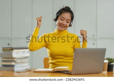 Happy Asian female student wearing wireless headphones studying online, listening to music, dancing, browsing websites, searching for information, chatting online on laptop in business study office.