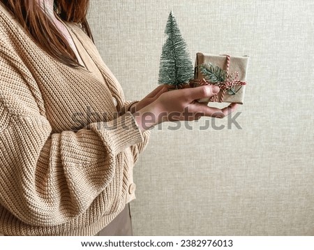 An unrecognizable brunette girl in a beige sweater holds a Christmas gift in eco packaging and a green Christmas tree in her hands. a small gift in honor of Christmas, new year.