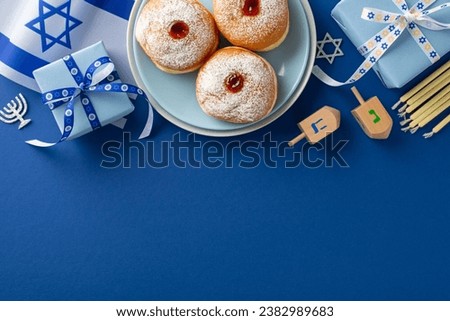 Partake in Hanukkah customs through a top-view picture of sufganiyot, Israeli flag, gift box with bow, candles, and dreidel on a blue background, with space for text or promotional material Royalty-Free Stock Photo #2382989683