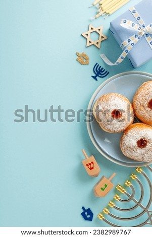Hanukkah holiday meal arrangement. Top view vertical image of Jewish sufganiyot on plates, Star of David, gift box, menorah candle holder, dreidel set on a pastel blue background with text or ad space