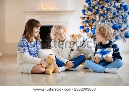 Christmas at home. Kids and dog under Xmas tree. Little boy and girl hug pet in Santa hat and open Christmas presents. Children play with animal. Winter holiday celebration. Blue and white theme.