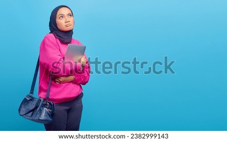 Photo of sad asian woman wearing jacket carrying laptop looking at empty space isolated on blue background