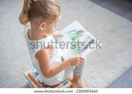 Little girl is sitting on stack of children's books and leafing through a book with pictures of fairy tales. High quality photo