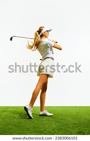 Elegant young girl with blonde hair, in stylish white clothes playing golf, swinging club on grass field isolated over white background. Concept of sport, hobby, beauty and fashion, relaxation, game