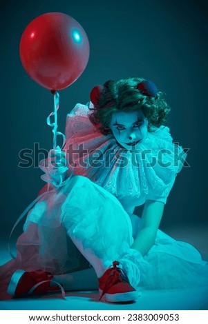 Mysterious old circus. A clown girl in a white circus dress sits on a dark stage, shrouded in haze, and looks intently at the balloon. Halloween.