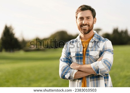 Portrait of smiling handsome middle aged man farmer wearing s stylish t shirt holding arms crossed looking at camera standing in green field, copy space. Successful business concept  