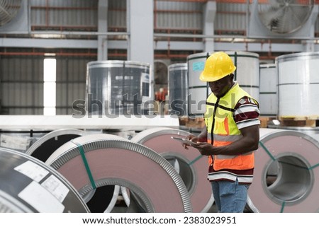 Metalwork manufacturing, warehouse of raw materials. Male factory worker inspecting quality rolls of metal sheet in aluminum material warehouse, wearing safety uniform and uses digital tablet