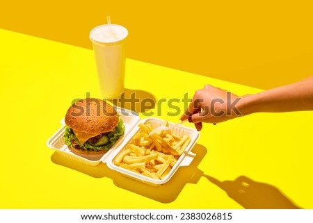 Unpacking tasting fast-food. Unrecognizable woman eat hamburger with soda and fried potato in paper boxes against vivid yellow background. Concept of junk food, menu, delivery, catering, take away. Ad