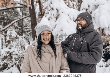 Young couple having fun in winter falling snow season in city park covered green trees pine, holiday vacation weekend, enjoying spending time together laughing smiling