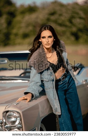 A beautiful woman stands next to a convertible in a field. Dressed in jeans and a jacket with fur. Beautiful and stylish look.