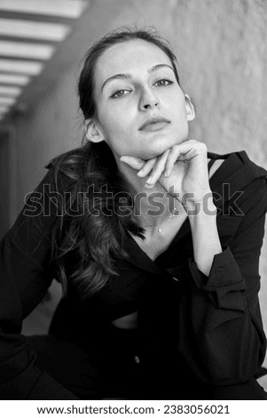 Black and white portrait. A beautiful young girl in a black shirt and black trousers is sitting on a chair. Casual look.
