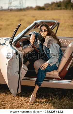A beautiful woman sits on a convertible in a field wearing sunglasses. Dressed in jeans and a fur jacket. Beautiful and stylish look.