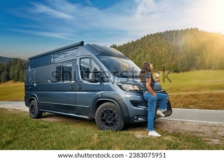 Happy, smiling attractive dark haired woman on a hike, leaning against a camper on a mountain road surrounded by mountains. Sunny day, adventure in the Dolomites, Italy. Royalty-Free Stock Photo #2383075951