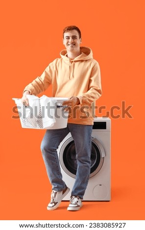 Young man with laundry near washing machine on orange background Royalty-Free Stock Photo #2383085927
