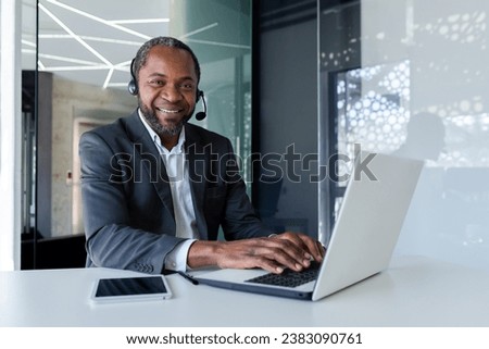 Portrait of african american businessman inside office at workplace, mature man using headset phone and laptop for remote consultation with clients, smiling and looking at camera.