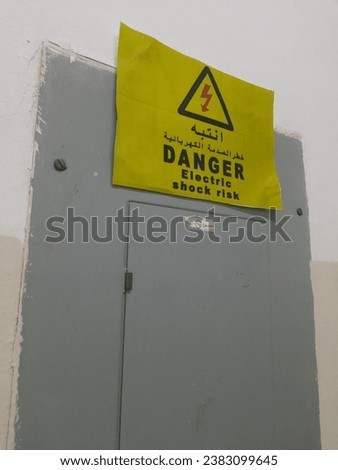 Egypt, Kafr El-Sheikh, 10-21-2023, the phrase “danger, electric shock risk” in Arabic and English on a yellow sticker on a gray electrical switch box attached to the wall, no smoking sign in arabic