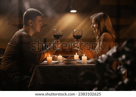 Trough window view on young couple in love drinking wine, celebrating Valentines day dining at home, having romantic dinner date with candles. Copy space Royalty-Free Stock Photo #2383102625