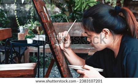 Asian woman painter creating art use a paintbrush to draw lettering designs on a wooden coffee shop sign. outdoor activities, People doing activities.