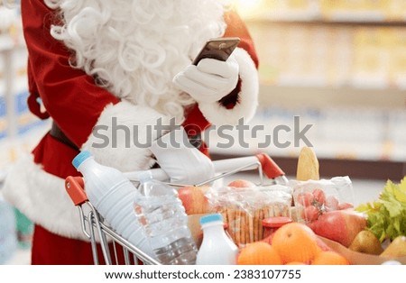 Santa Claus doing grocery shopping at the supermarket, he is pushing a trolley full of groceries and using a smartphone, hands close up Royalty-Free Stock Photo #2383107755