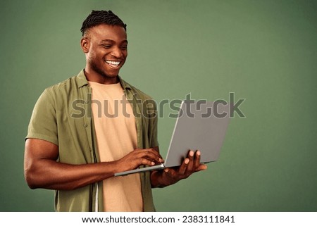 Happy african american freelancer in casual khaki shirt looking on laptop screen while typing on keyboard. Positive handsome man standing over green background and surfing internet.