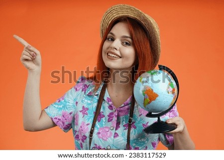 Close up of woman holding small globe and pointing at the empty area for advertisement smiling, isolated on orange background. Portrait of female traveler. Royalty-Free Stock Photo #2383117529