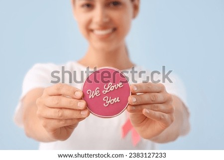 Young woman holding cookie with text WE LOVE YOU on light background, closeup. Breast cancer awareness concept