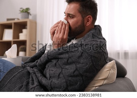 Sick man wrapped in blanket with tissue blowing nose on sofa at home. Cold symptoms Royalty-Free Stock Photo #2383138981