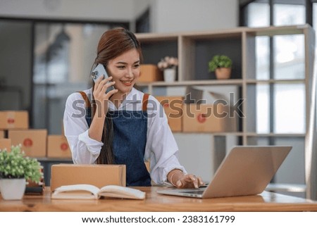 A portrait of a young Asian woman, e-commerce employee sitting in the office full of packages in the background write note of orders and use smartphone, for SME business ecommerce and delivery