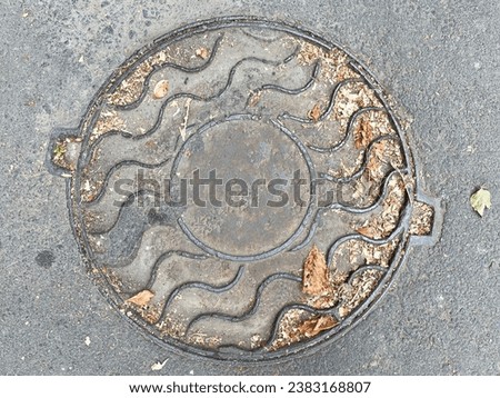 A round metal hatch with wavy lines on the surface is built into one level with gray textured asphalt, close-up, top view. There are small particles of dry brown leaves on the surface of the hatch.